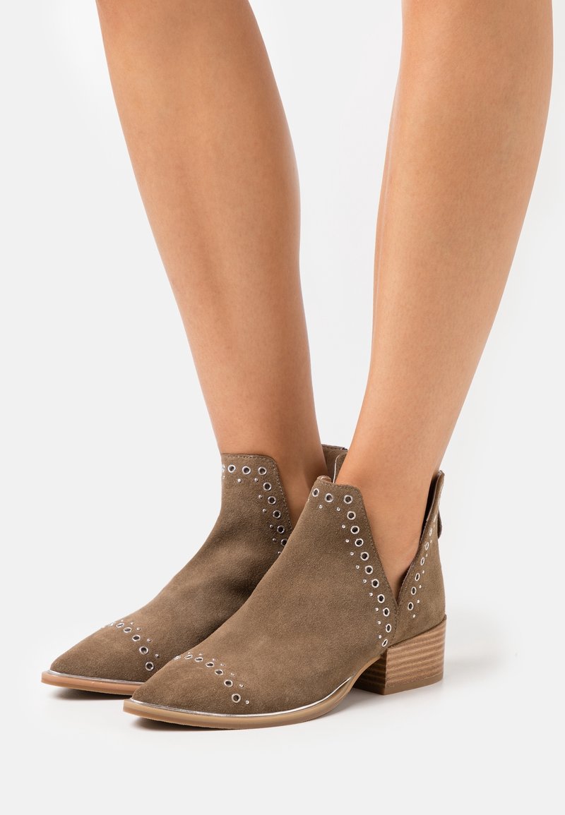 Bottines en daim marron à œillets argentés, bouts pointus et talons en bois empilés basportées sur jambes nues.