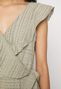 Světle olivově zelené šaty s výstřihem do V, volánkovými rukávy, texturovanou tkaninou a malými výřezovými vzory po celém povrchu.