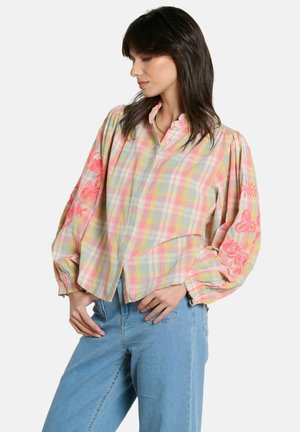 Frau trägt pastellfarbenes kariertes Bluse mit rosa Blumenstickerei auf den gepufften Ärmeln und hellblauen Jeans, posiert mit den Händen in der Hüfte.