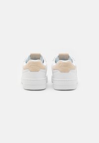 Baskets blanches avec un dessus en cuir lisse, des accents beiges au niveau du talon et une semelle en caoutchouc texturée. Design minimaliste avec un détail de marque emblématique.