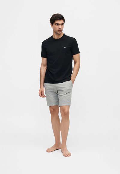 Νέος άνδρας που στέκεται ξυπόλητος, φοράει μαύρο μπλουζάκι και γκρι σορτς, με το ένα χέρι στην τσέπη, κοιτάζοντας προς το πλάι σε απλό φόντο.