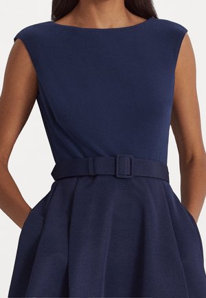 Frau trägt ein ärmelloses marineblaues Kleid mit einem U-Boot-Ausschnitt und einem passenden Gürtel in der Taille, Hände in den Taschen.