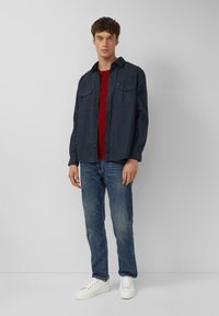 Dunkelblaues Hemd über einem roten Pullover, kombiniert mit hellblauen Jeans und weißen Sneakers. Das Hemd hat zwei Brusttaschen.