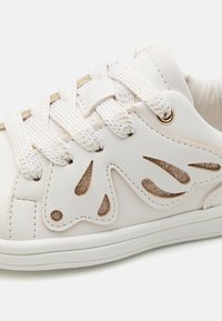 Witte synthetische sneaker met decoratieve uitsparingen, gouden accenten en gestructureerde veters. Heeft een gladde afwerking en een platte rubberen zool.