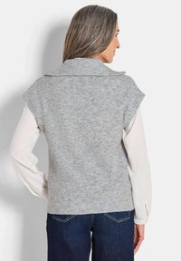 Gilet en maille grise avec une texture côtelée et un col large, porté par-dessus une chemise blanche à manches longues, associé à un jean bleu foncé.