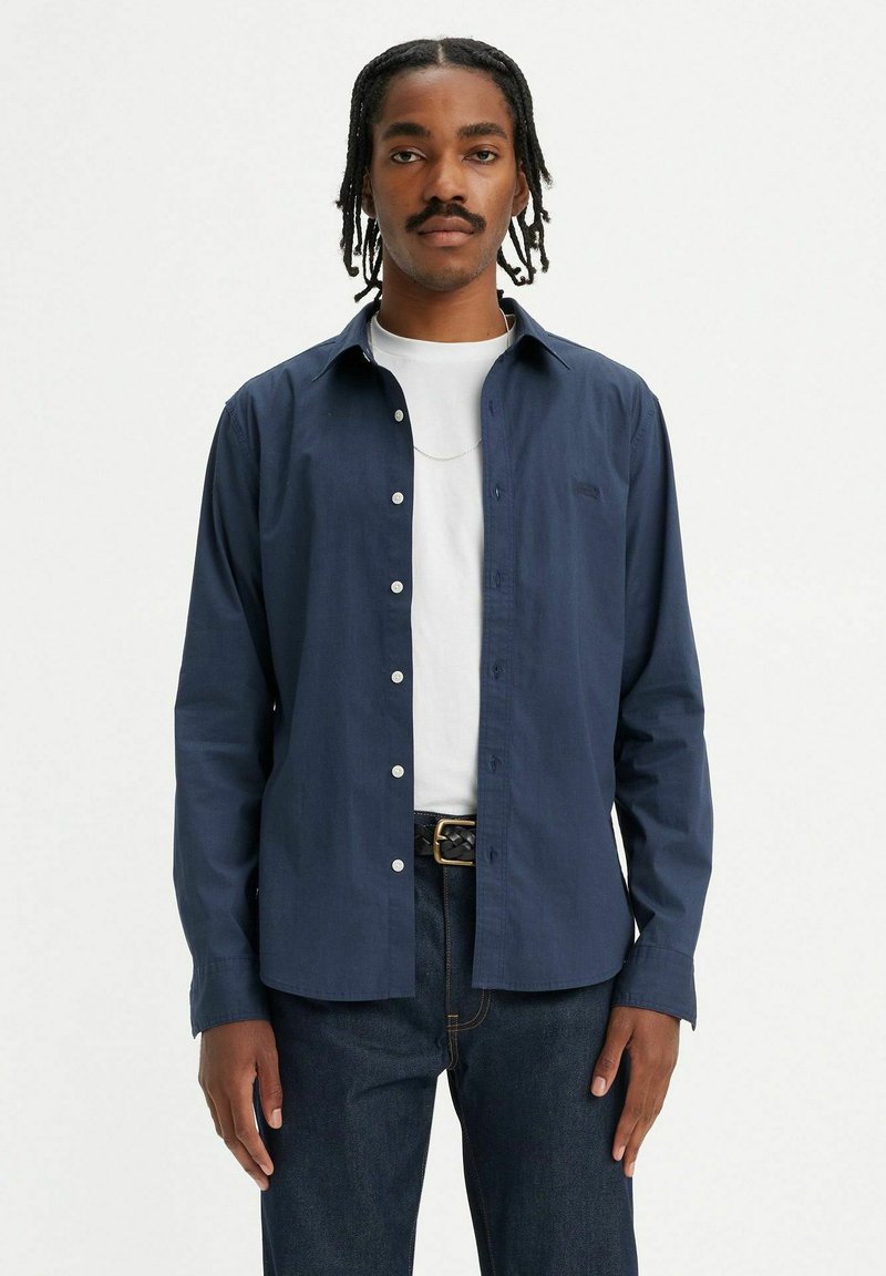 Camicia a maniche lunghe blu navy con colletto button-down, in cotone, e bottoni bianchi. Indossata sopra una t-shirt bianca e abbinata a jeans scuri.