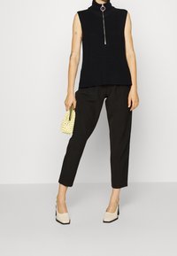 Černý bezrukávový pletený rolák se stříbrným zipem, doplněný černými kalhotami a béžovými botami na blokovém podpatku. Drží malou žlutou tašku.