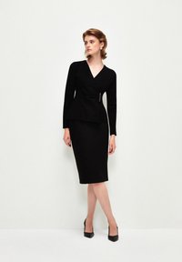 Vestido negro envolvente con mangas largas, cintura peplum y un detalle plateado. Tela suave, corte hasta la rodilla, combinado con tacones negros.