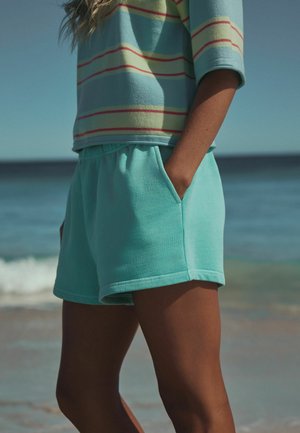 Isik, kes kannab türkiissiniseid lühikesi pükse ja triibulist sinise ja roosa tooniga tuba, seisab randal käsi taskus.