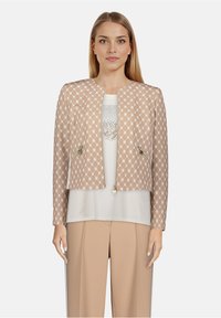 Veste courte beige avec motif en losange, ferrures dorées et deux poches avant, portée sur un haut clair et un pantalon beige.