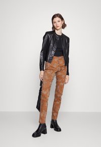 Jaqueta de imitação de couro preta com ombros texturizados, combinada com calças castanhas estampadas e botas de salto pretas. Modelo posa sobre um fundo neutro.