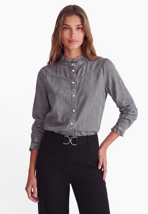 Vrouw draagt een grijze blouse met knopen en ruches aan de kraag en manchetten, gecombineerd met een zwarte broek en een zwarte riem met zilveren gesp.