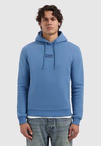 Man met krullend haar, gekleed in een blauwe "PURE PATH" hoodie en versleten blauwe jeans, staand voor een effen lichtgrijze achtergrond.