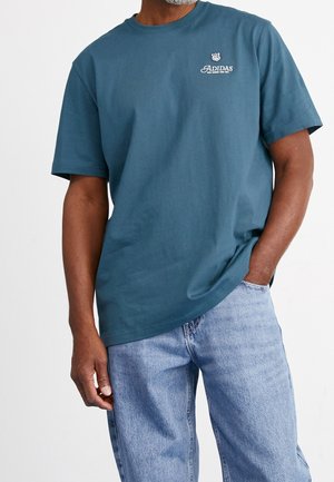 Morska zielona bawełniana koszulka z okrągłym dekoltem i krótkimi rękawami. Na piersi znajduje się białe logo Adidas. W zestawieniu z jasnoniebieskimi dżinsami.