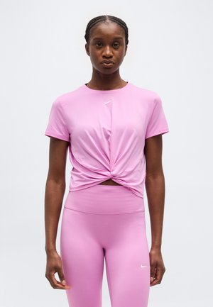 Jeune femme aux cheveux tressés portant un haut de sport rose avec un devant torsadé et un legging taille haute assorti, se tenant devant un fond uni.