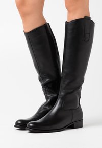 Bottes en cuir noir, montantes jusqu'au genou, avec un design épuré, des semelles plates et une fermeture éclair sur le côté pour un enfilage facile. Texture lisse ; accents minimalistes.