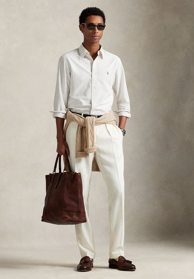 Camisa às riscas creme com botões, suéter bege amarrado à cintura, calças brancas, bolsa de couro escuro e mocassins escuros. Óculos de sol pretos completam o visual.