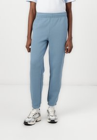 Lyseblå bomullssweatpants med elastisk midje, smalnende mansjetter og en teksturert logo detalj på forsiden. Kombinert med sølvfargede sneakers.