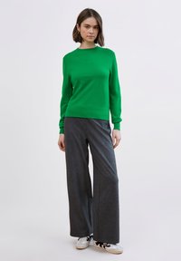 Zelený pletený svetr s dlouhými rukávy, doplněný šedými pruhovanými širokými kalhotami a bílými teniskami s černými detaily.