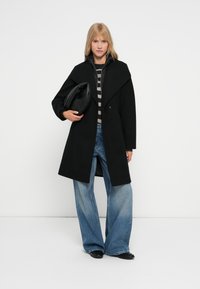 Cappotto di lana nero con colletto alto, sovrapposto a una maglia a righe grigie, abbinato a jeans blu a gamba larga e una borsa nera.