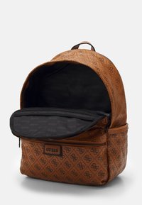 Ruskea Guess-reppu, jossa on monogrammikuvio, etu-zip-tasku ja osittain avoin pääosasto, josta näkyy musta sisävuori, jossa on brändin nimi.