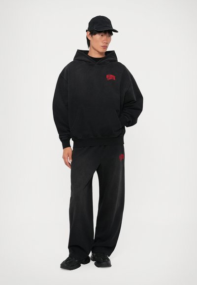 Sudadera de forro polar negra con un bolsillo frontal y un logo rojo, combinada con pantalones de forro polar negros que tienen un pequeño logo rojo, y zapatos deportivos negros.