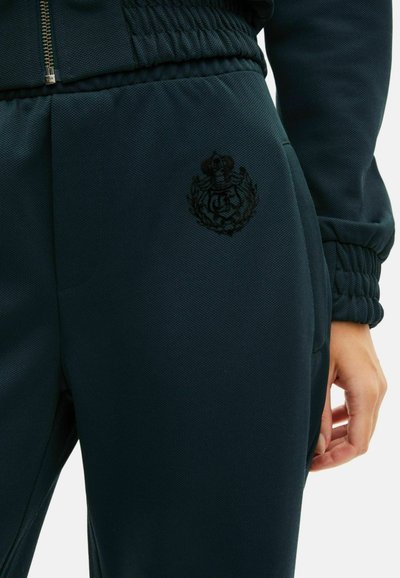 Mørkegrønne joggingbukser lavet af struktureret stof, med elastisk liv, og broderet emblem i sort på låret.