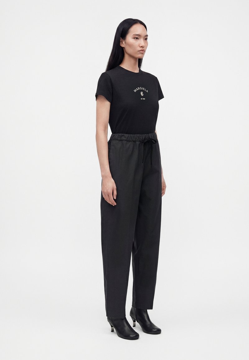 Femme aux longs cheveux noirs portant un t-shirt noir à manches courtes, un pantalon noir taille haute et des bottines noires à talons, debout de côté.