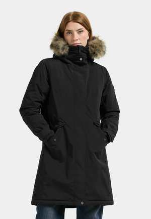 Femme portant un long manteau d'hiver noir avec une capuche bordée de fourrure, les mains dans les poches, debout devant un fond clair uni.