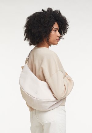 Vrouw met krullend haar die een beige sweatshirt en witte broek draagt, met een grote off-white schoudertas op haar rug, zijwaarts kijkend.