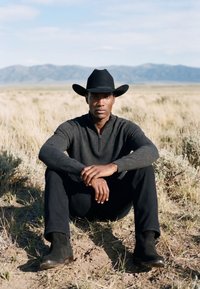 Homme portant un chapeau de cowboy noir, un pull foncé, un pantalon et des bottes, assis en tailleur sur une prairie sèche avec des montagnes en arrière-plan.