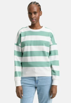 Vrouw met gevlochten haar, die een witte en muntgroene horizontaal gestreepte trui en lichtblauwe spijkerbroek draagt, staand tegen een witte achtergrond.