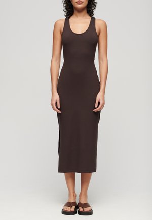 Femme portant une robe ajustée sans manches, marron foncé, longueur cheville, et des sandales marron, debout devant un fond clair uni.