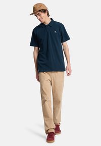Marineblå polo t-shirt med korte ærmer, et lille logo, parret med lysebrune kordfløjsbukser og røde sneakers, stylet med en brun kasket.