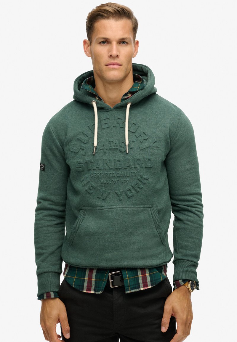 Sudadera verde con logo texturizado, capucha con cordón y bolsillo frontal, superpuesta a una camisa de cuadros. Tejido suave, diseño ajustado.