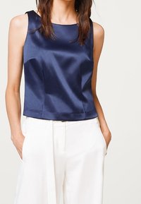 Navy blue satin sleeveless top with a round neckline and straight cut, paired with white high-waisted trousers. Smooth texture, minimal design.