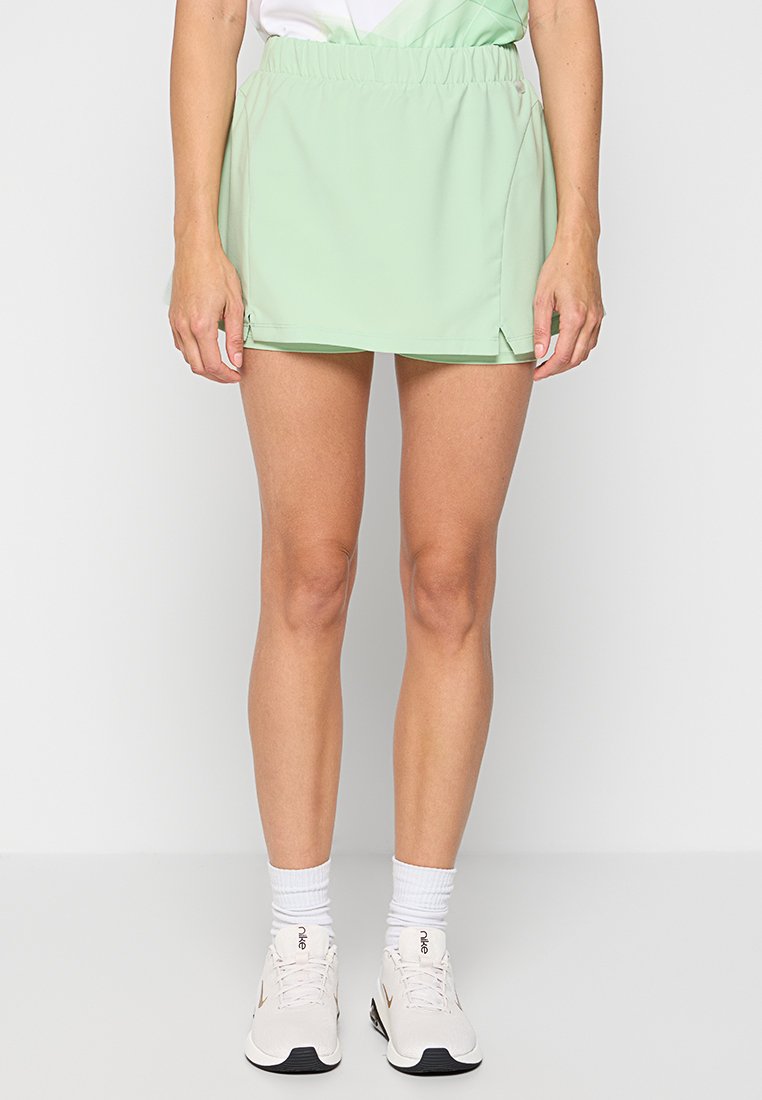 Falda atlética de color verde claro con cintura elástica, aberturas laterales y una textura suave. Llevada con calcetines blancos y zapatillas de running en tonos neutros.