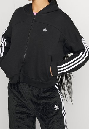 Personne portant un sweat à capuche Adidas noir zippé avec des bandes blanches sur les manches et un pantalon Adidas noir assorti avec des bandes blanches.