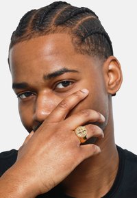 Bague sigillaire en or avec un visage rond présentant un design en relief, portée sur une main placée près du visage, avec des cheveux tressés visibles.