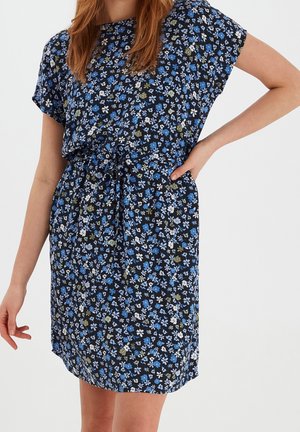Kvinde iført sort kjole med korte ærmer med små blomsterprint i blå, hvid og grøn og bindebånd i taljen, stående mod en ensfarvet lys baggrund.