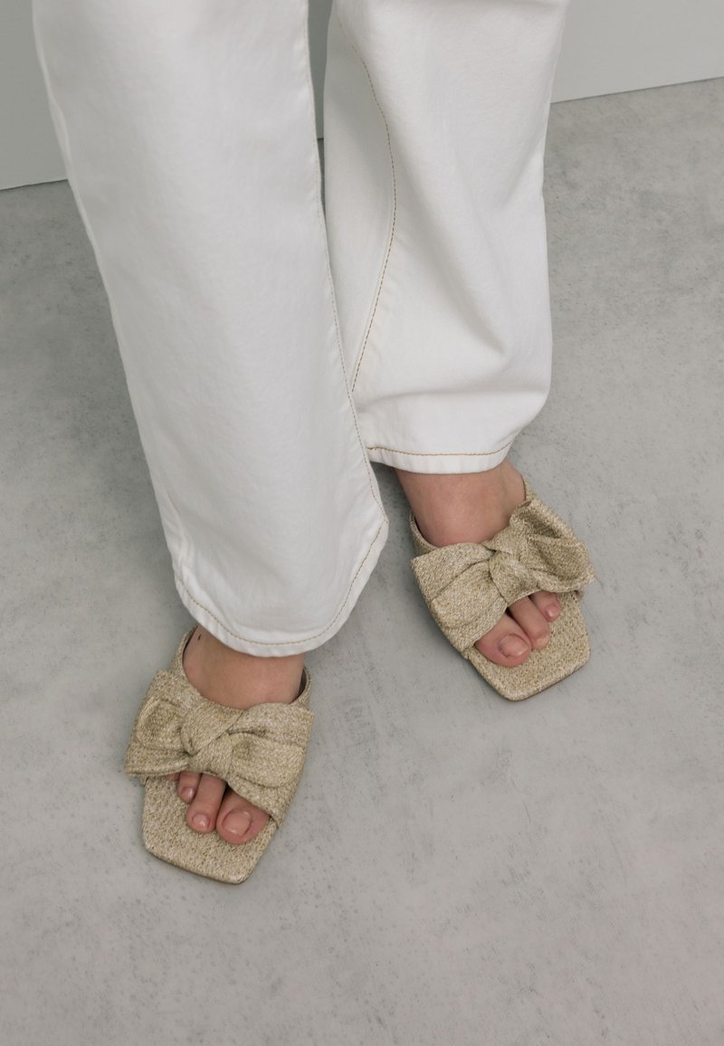 Gouden gevlochten slide-sandalen met een grote strikdetail aan de voorkant. Vierkante tenen, natuurlijke textuur, gedragen met witte, losse broek.