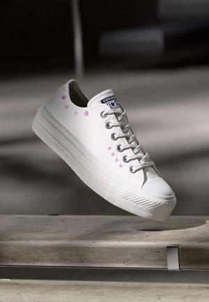Biały tenis Converse z różowymi kwiatowymi wzorami i haftem krzyżykowym unoszący się nad betonową powierzchnią w miękkim świetle.