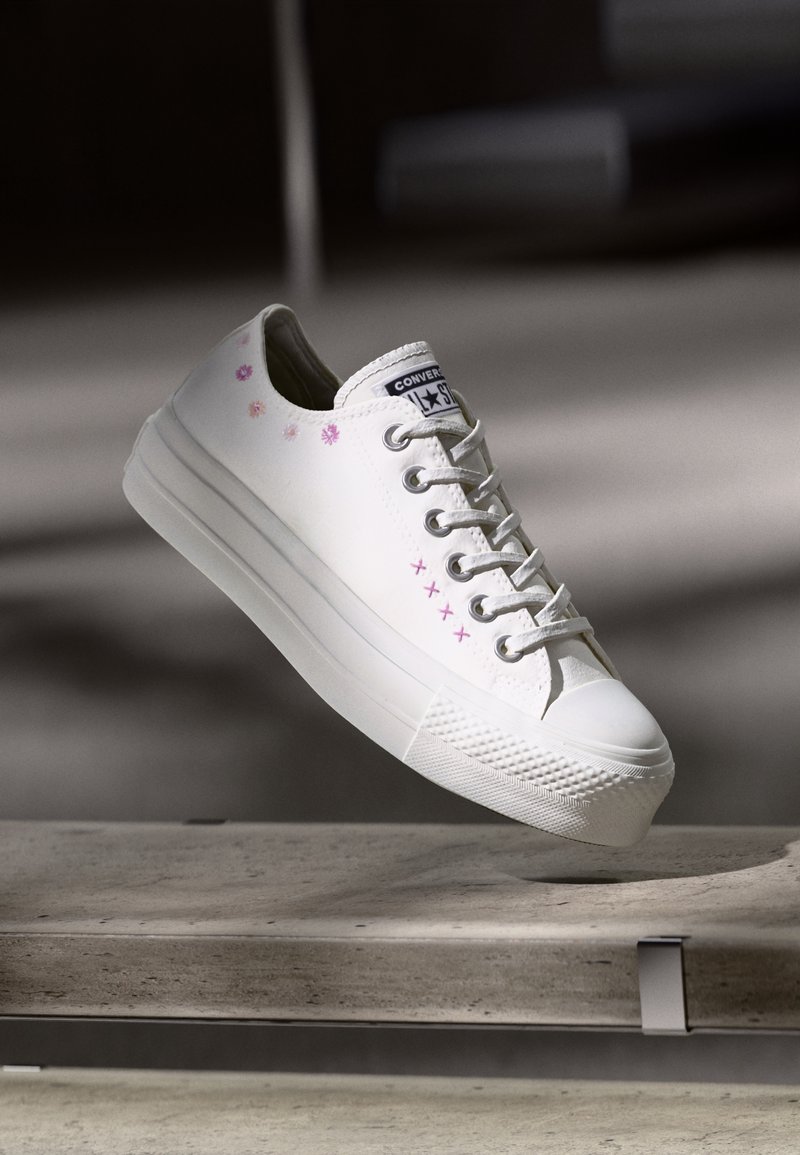 Baskets blanches Converse avec motifs floraux roses et broderies au point de croix flottant au-dessus d'une surface en béton dans une lumière douce.