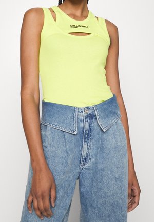 Kobieta nosząca bezrękawnik w neonowo żółtym kolorze z wycięciem oraz niebieskie dżinsy z wysokim stanem i szerokim złożonym paskiem.