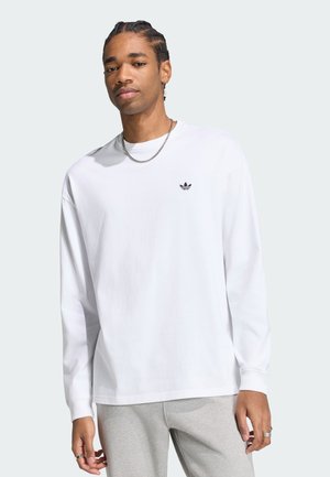 Mann mit geflochtenem Haar, der ein weißes langärmeliges Adidas-Shirt und graue Hosen trägt, steht vor einem schlichten hellen Hintergrund.