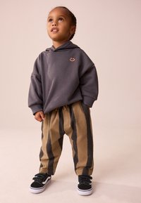 Grauer Kapuzenpullover mit einem Smiley-Logo, beige-schwarze gestreifte Hose, schwarze niedrige Sneakers mit weißen Sohlen. Weicher Stoff, lockere Passform.