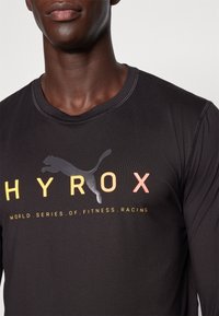 Maglietta nera a maniche lunghe con scollo tondo, caratterizzata da un testo colorato e dal logo Puma. Il materiale ha una texture liscia e atletica.