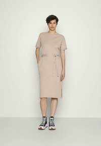 Vestido bege de manga curta com um cinto na cintura, bolsos laterais e um corte até ao joelho, combinado com ténis preto e branco.