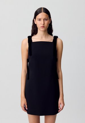Donna con lunghi capelli scuri che indossa un vestito nero senza maniche con bretelle larghe e scollatura rettangolare, su uno sfondo piatto.