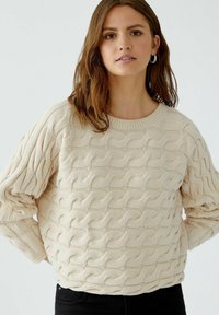 Beige gebreide trui met kabelpatroon, ronde halslijn en verlaagde schouders, gecombineerd met zwarte jeans. Soepele textuur en ontspannen pasvorm.